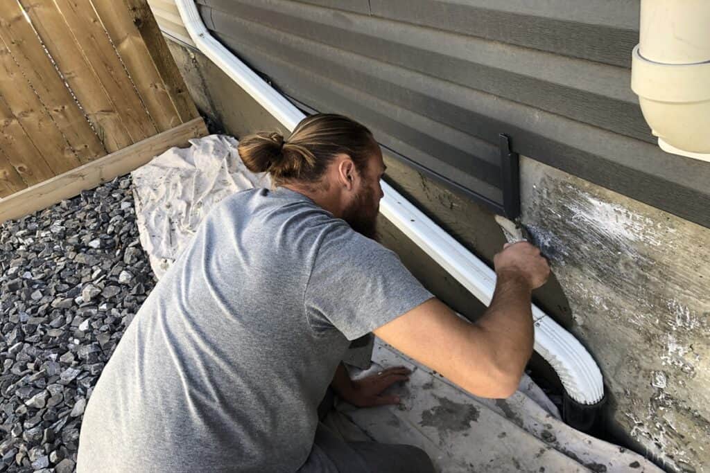 Parging repair on house foundation in Edmonton A contractor in a gray shirt applies fresh parging to a home's foundation using a trowel. The surface is being repaired near a downspout, with a protective drop cloth covering the ground.