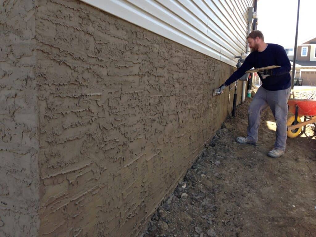 Parging application on house exterior in Edmonton A contractor in work gloves and a navy shirt applies a textured parging coat to a house foundation using a trowel. The fresh parging has a rough, layered finish, protecting the concrete foundation beneath. A wheelbarrow and construction tools are in the background.