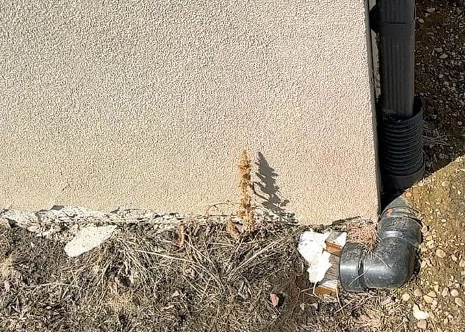 Acrylic stucco deterioration where the wall finish meets soil near a downspout in Edmonton